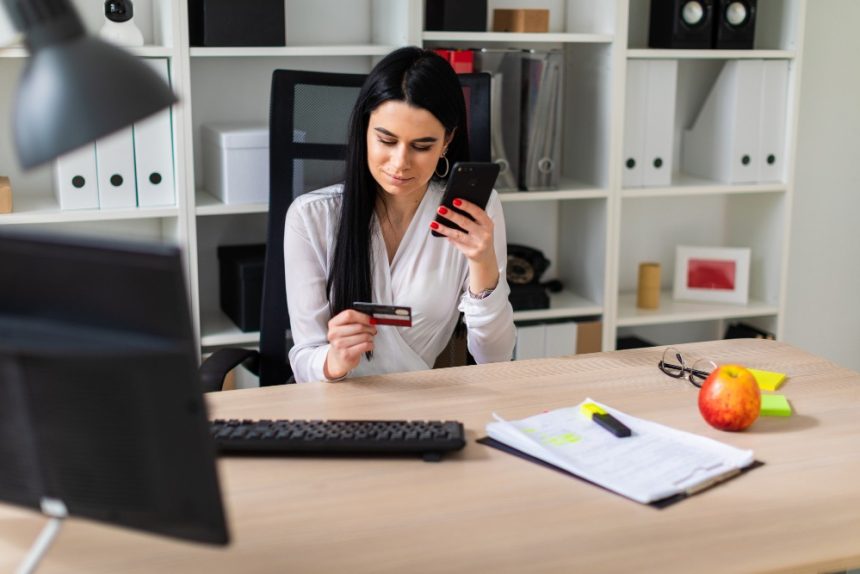 devojka za radnim stolom u ruci drzi telefon i platnu karticu
